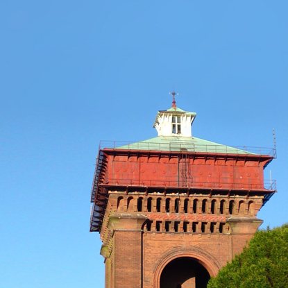 The top part of Jumbo Water Tower is seen against a blue sky. The building has four stacks of red coloured bricks which form the legs of the tower and are separated by arches. Two layers of brick alcoves sit on top of the legs. There are metal railings surrounding a huge square red metal water tank. There is a green pitched roof topped by a small white square room with a window.
